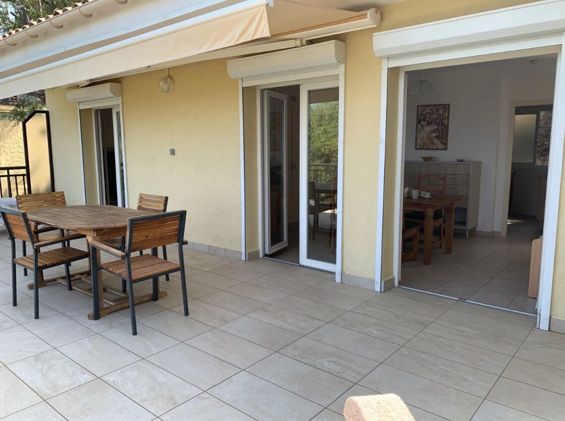 An outdoor patio area with a wooden table and chairs, a covered seating area, and sliding glass doors leading to an interior space.