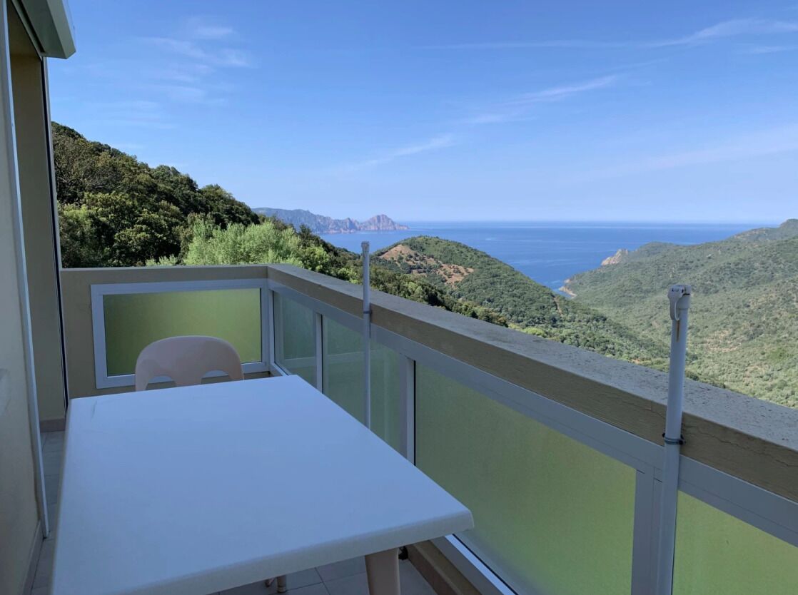 Un balcon avec une table et une chaise, donnant sur un paysage côtier avec des collines et la mer.