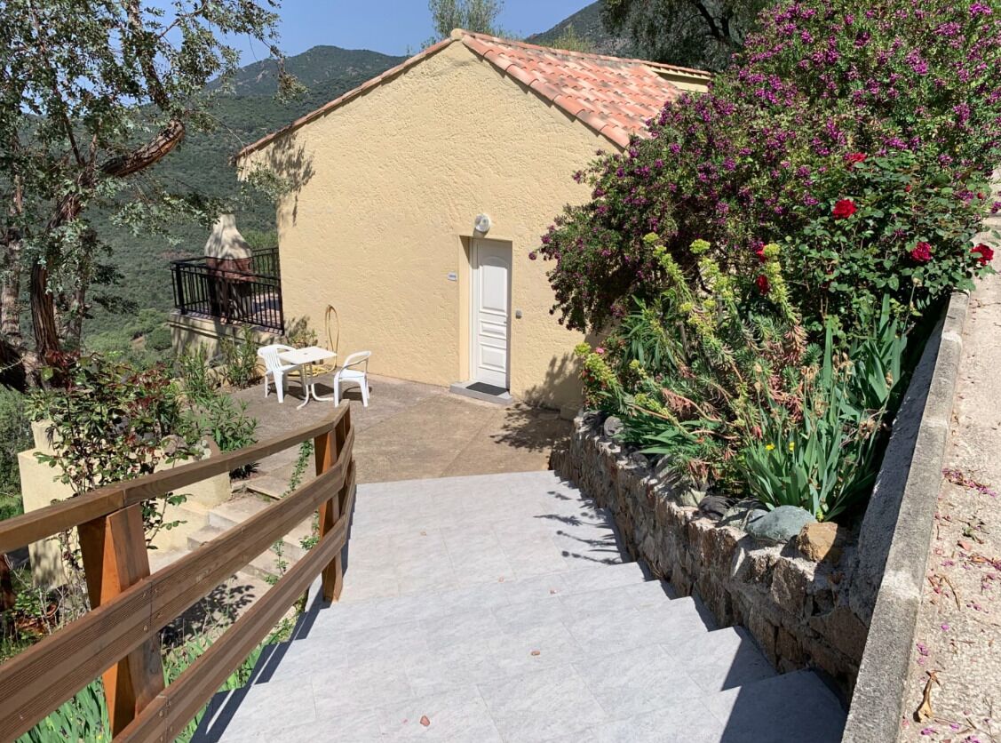 Un bâtiment avec un toit recouvert de tuiles, une porte blanche et un petit balcon avec une chaise. Au premier plan, on aperçoit une clôture en bois et, à droite, un mur de pierre orné de plantes et de fleurs.