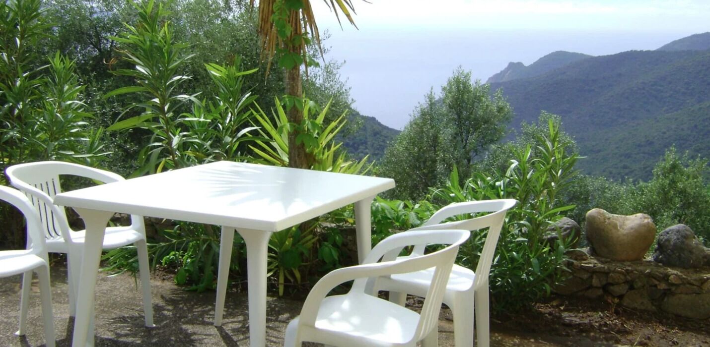 Une table et des chaises blanches sont disposées à l’extérieur, offrant une vue sur les montagnes et la verdure en arrière-plan.