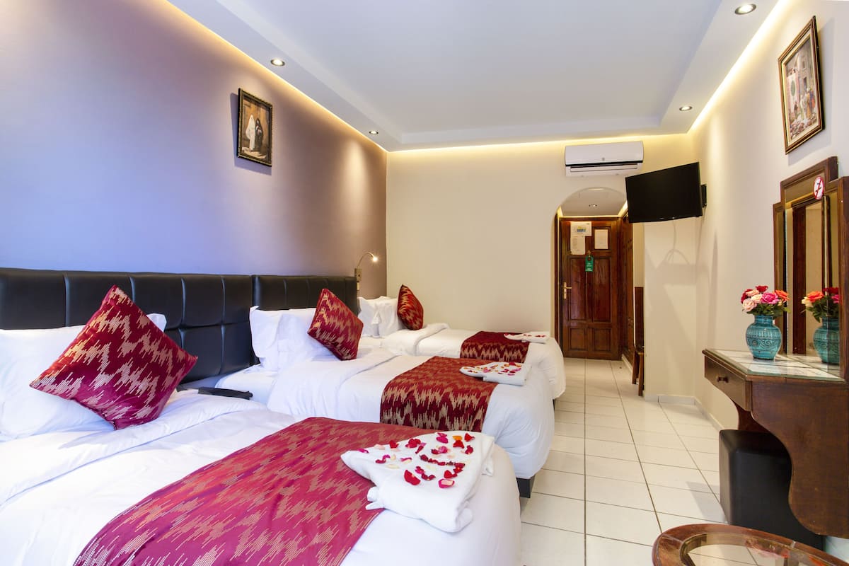 A hotel room with three beds, each with white linens and red patterned pillows. There is a wooden desk with a vase of flowers, and a television mounted on the wall.