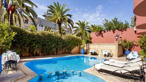 Piscina con sillones y mesas, rodeada de palmeras y un muro de piedra.