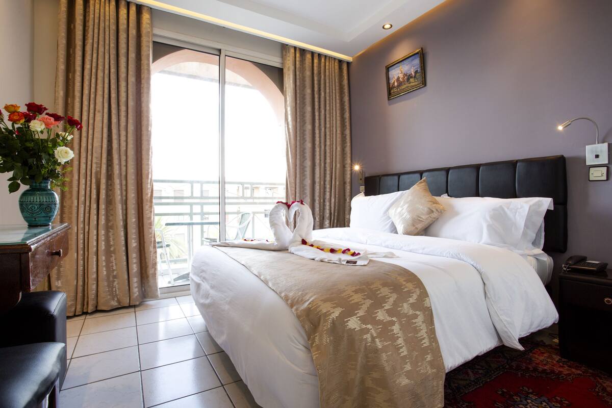 A hotel room with a large bed, a vase of flowers, a nightstand, and a view of the outdoors.