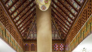 Un comedor con mesas dispuestas para dos, con camino de mesa azul, servilletas blancas y cristalería transparente. La habitación tiene un techo alto y ornamentado con diseños intrincados y una columna central imponente.