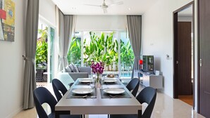 A modern dining area with a table set for four, a ceiling fan, and a view of a lush garden through large windows. - Tropical Stay at 2BR Pool Villa Greens 8 (Rawai)