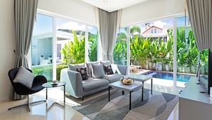 A modern living room with a grey sofa, a black chair, a coffee table, and a TV. There is a large window with a view of a swimming pool and greenery. - Tropical Stay at 2BR Pool Villa Greens 8 (Rawai)