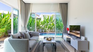 A modern living room with a sofa, coffee table, and TV, overlooking a swimming pool and lush greenery. - Tropical Stay at 2BR Pool Villa Greens 8 (Rawai)