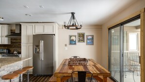 Eine Küche mit weißen Schränken, einem Edelstahlkühlschrank, einem Holztisch mit Stühlen und einem großen Fenster mit Blick nach draußen.