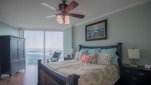 A bedroom with a large bed, a ceiling fan, a nightstand, a dresser, and a view of the ocean.