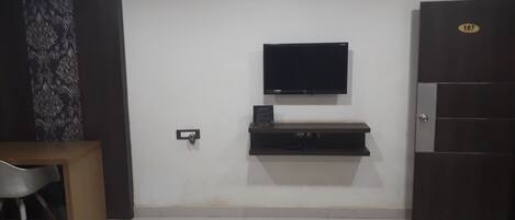 A room with a flat-screen TV mounted on the wall, a dark wooden door with a number plate, and a small table with a chair.