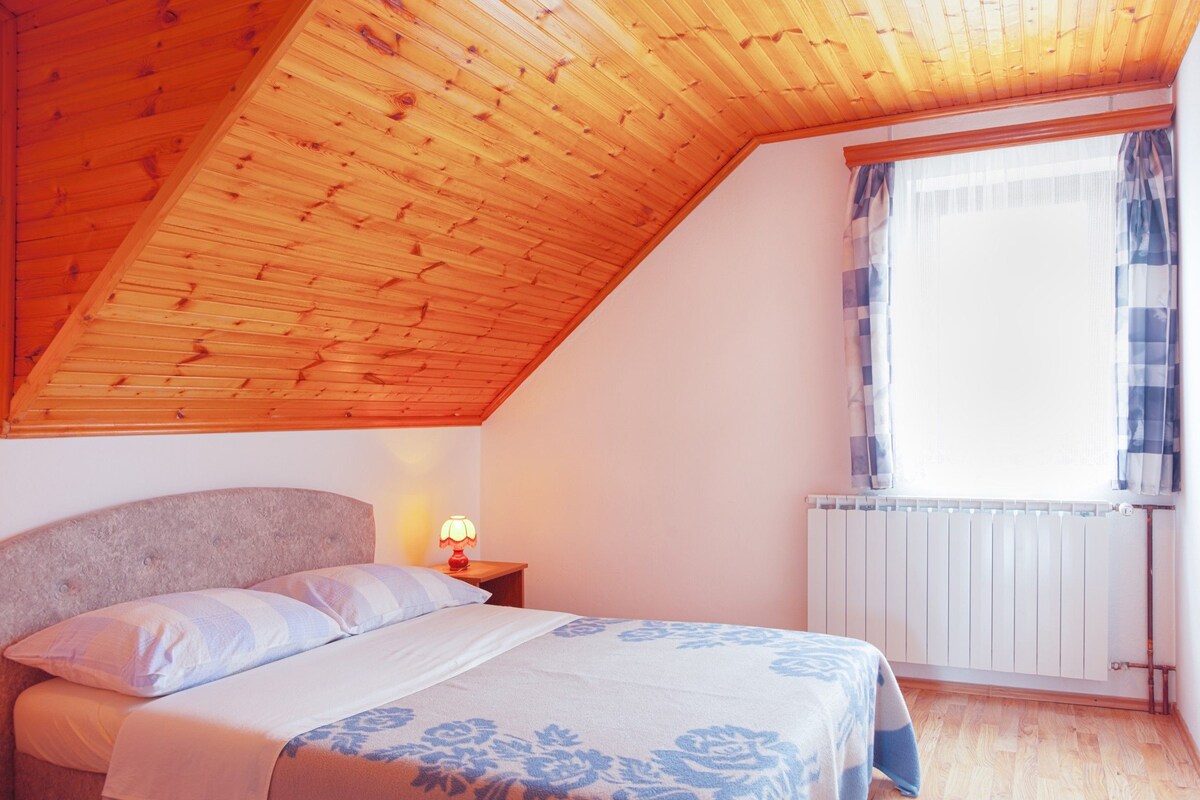A bedroom with a sloped ceiling, a bed with a headboard, a bedside lamp, and a window with curtains.
