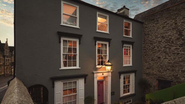 A multi-story building with white-framed windows, a central entrance with a red door, and a stone wall on one side. - One Drake Road Hotel (Tavistock)