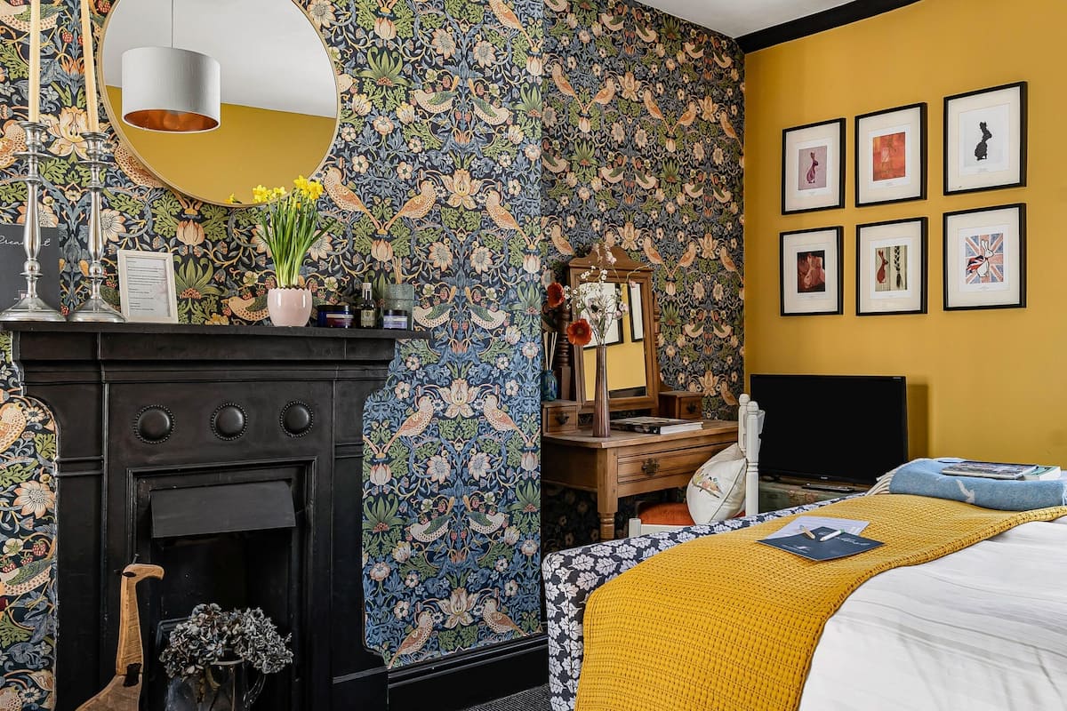 Une pièce avec une cheminée, un lit recouvert d’une couverture jaune, un bureau avec un ordinateur et un mur orné de tableaux.