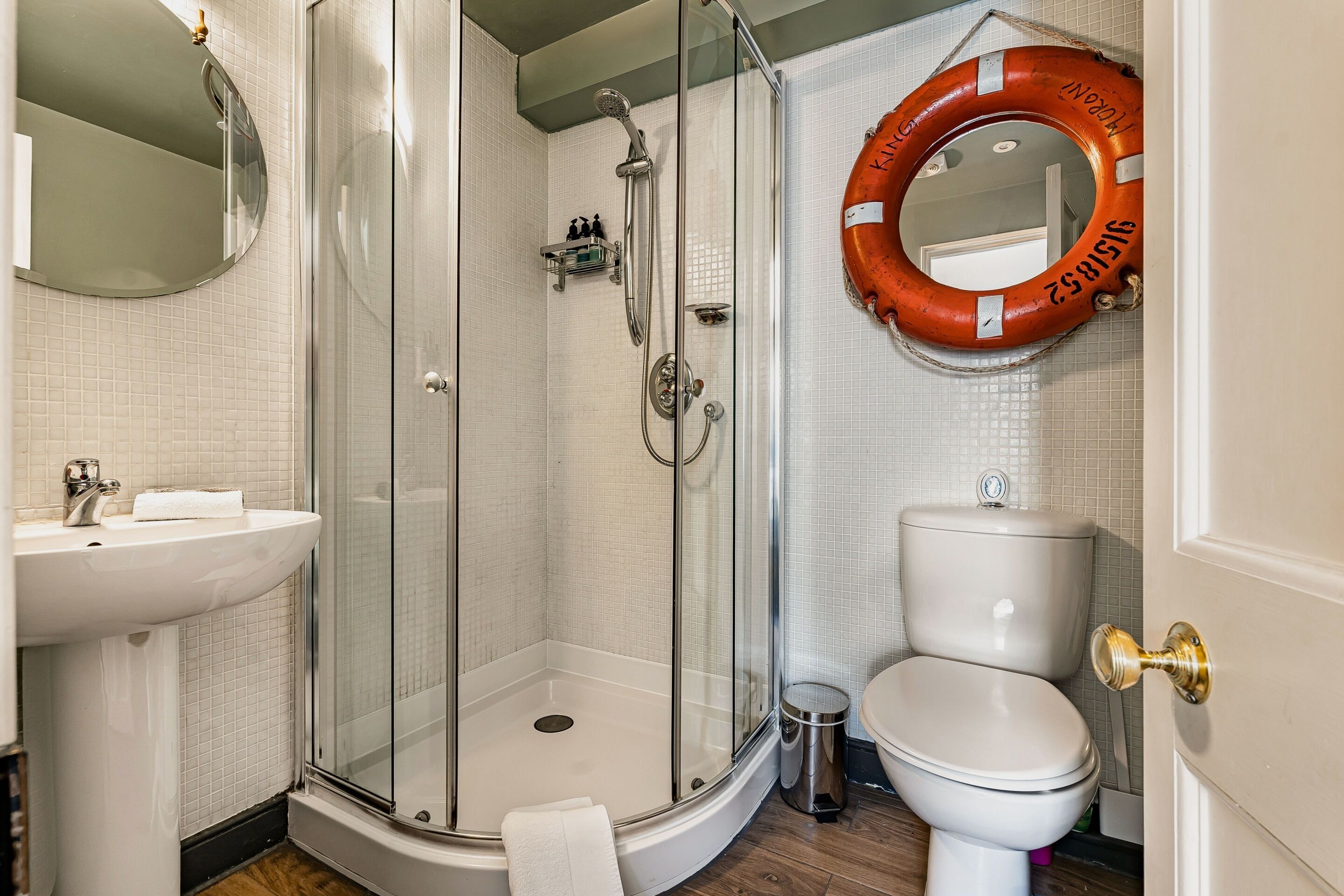 Un baño con ducha de vidrio, lavamanos blanco, inodoro y espejo redondo.