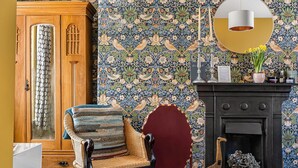 A room with a patterned wallpaper, a wooden cabinet, a wicker chair, a red upholstered chair, a fireplace, and a hanging light fixture. - One Drake Road Hotel (Tavistock)