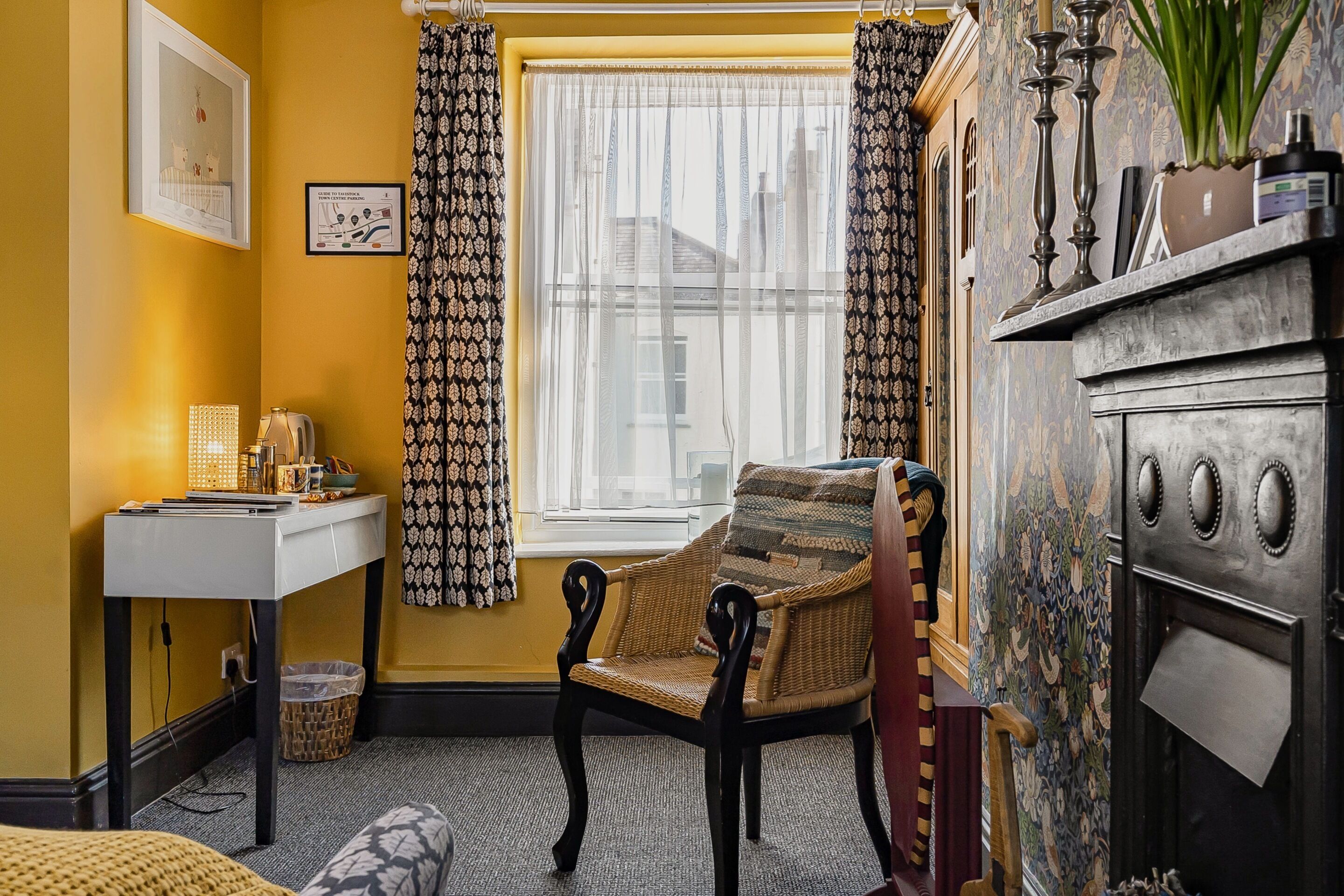 A room with a window, a chair, a small table, a fireplace, and a patterned wall.