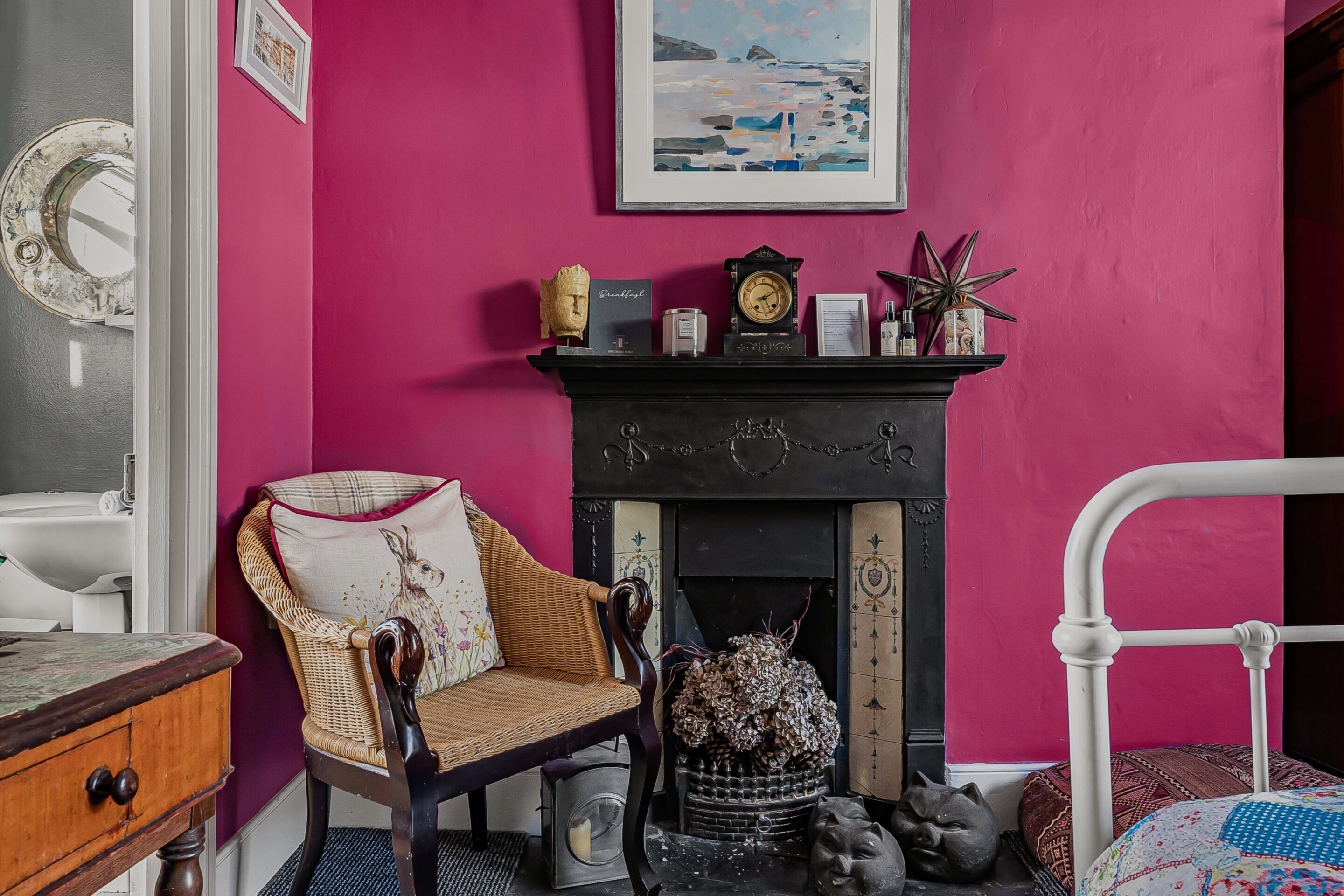A room with a pink wall, a fireplace, a wicker chair, a bed, and a white radiator.