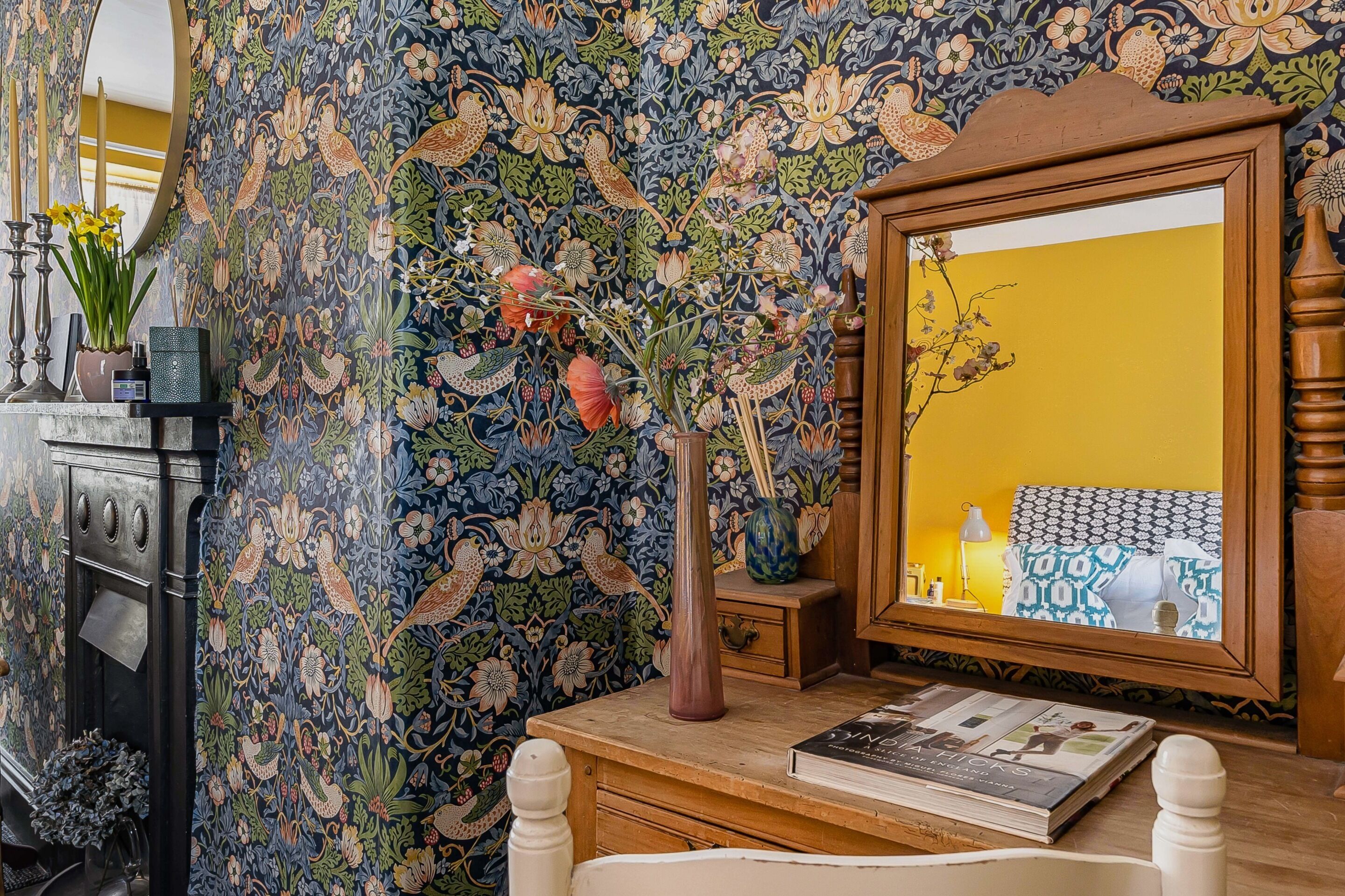 A room with a floral wallpaper, a wooden dresser, a mirror, and a book on the dresser.