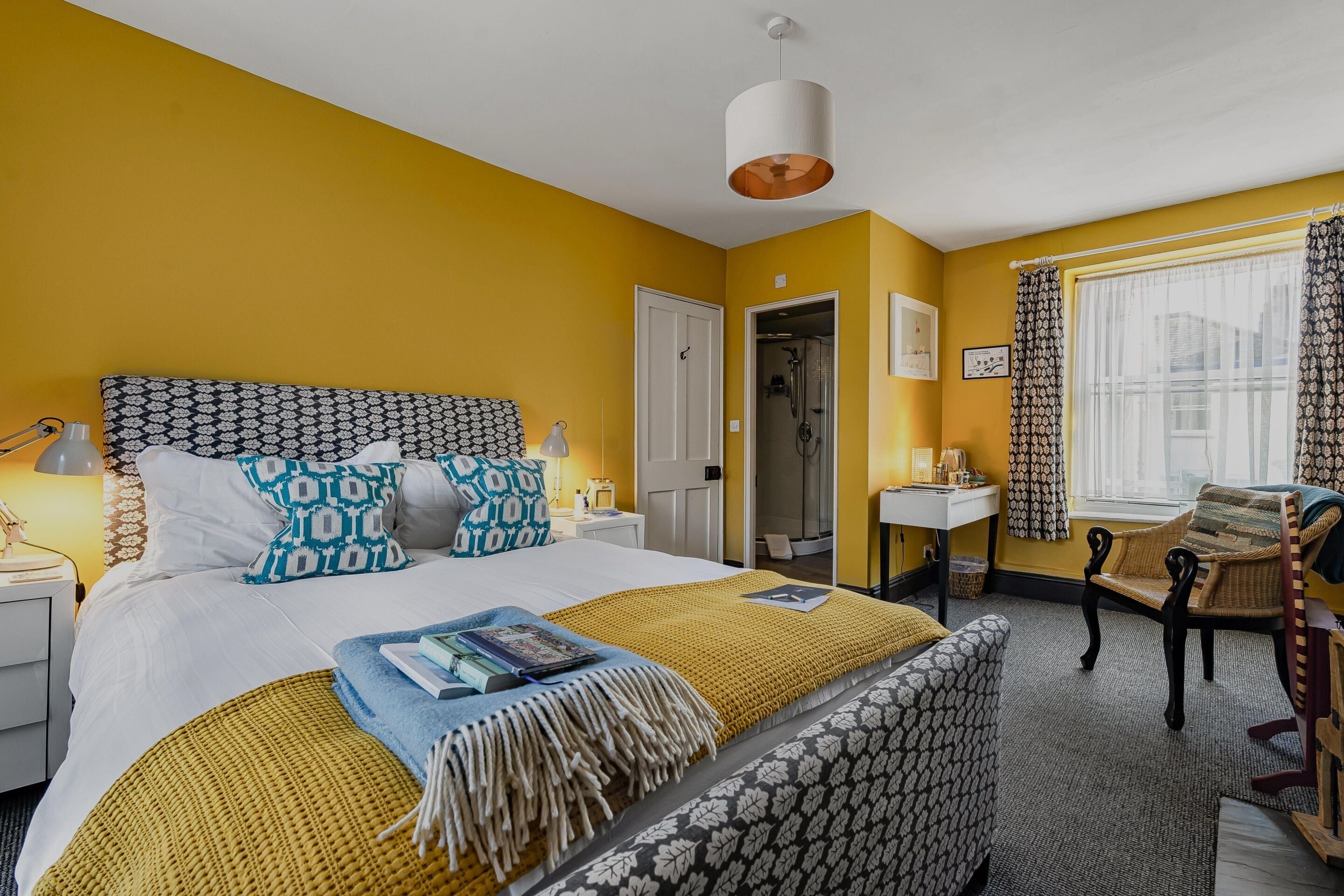 A hotel room with a bed, bedside tables, a desk, a chair, a window with curtains, and a door leading to a bathroom.