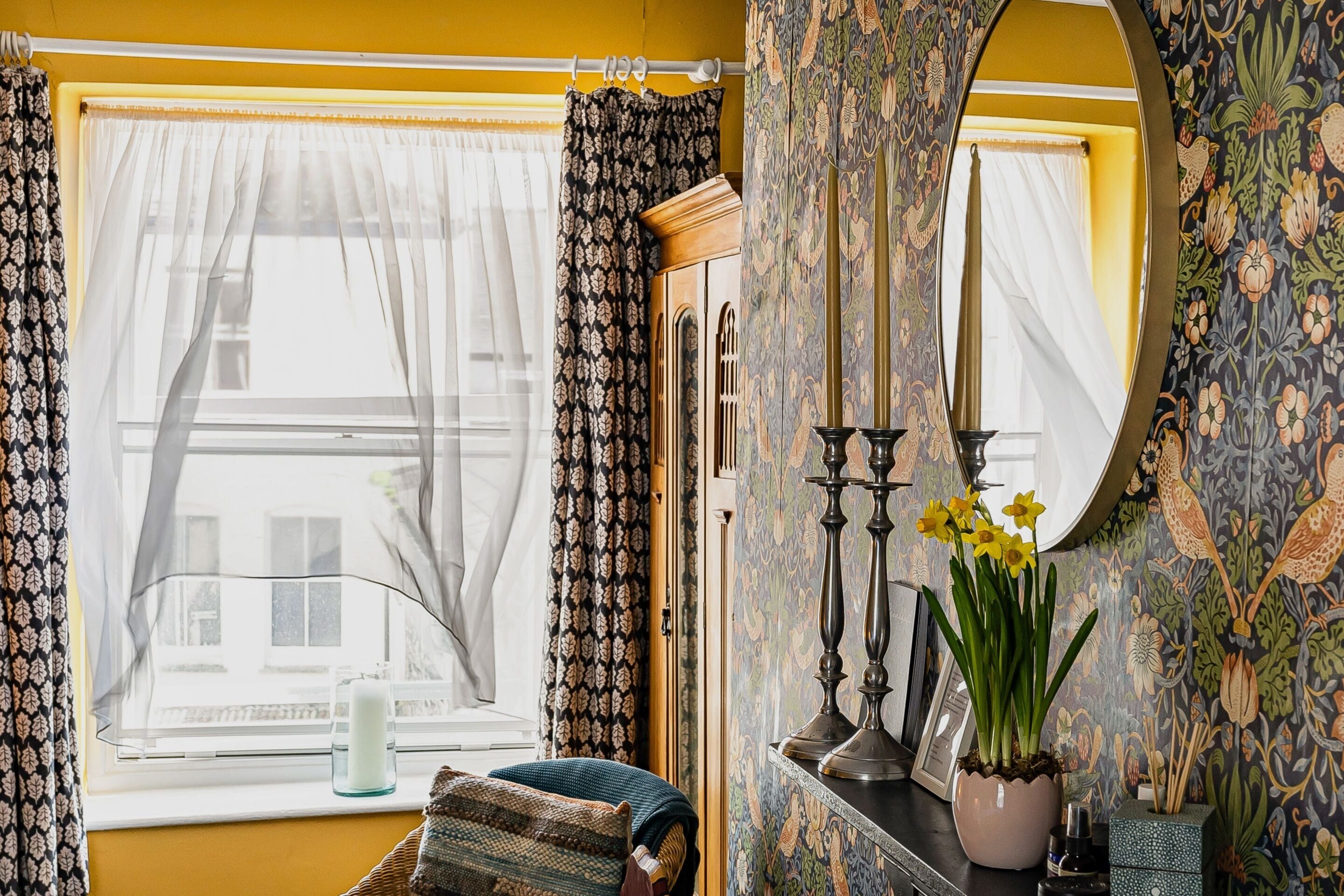 A room with a window, curtains, a mirror, a vase with flowers, and a candle holder.