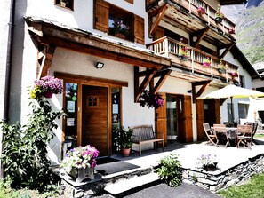 Una cabaña tradicional con balcones de madera, un patio con mobiliario exterior y un paisaje montañoso de fondo.