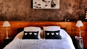 Un dormitorio con cabecera de madera, dos mesitas de noche con lámparas, cama con ropa de cama blanca y almohadones con estampado de corazones, y un cuadro colgado en la pared.