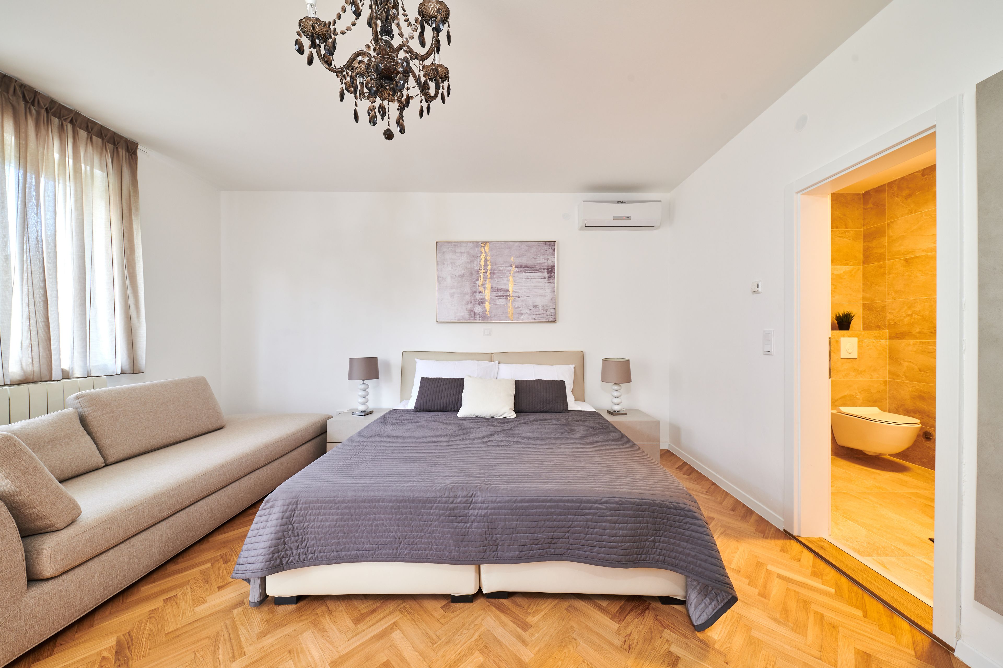 A modern bedroom with a large bed, a sofa, a chandelier, and a bathroom visible through an open door.