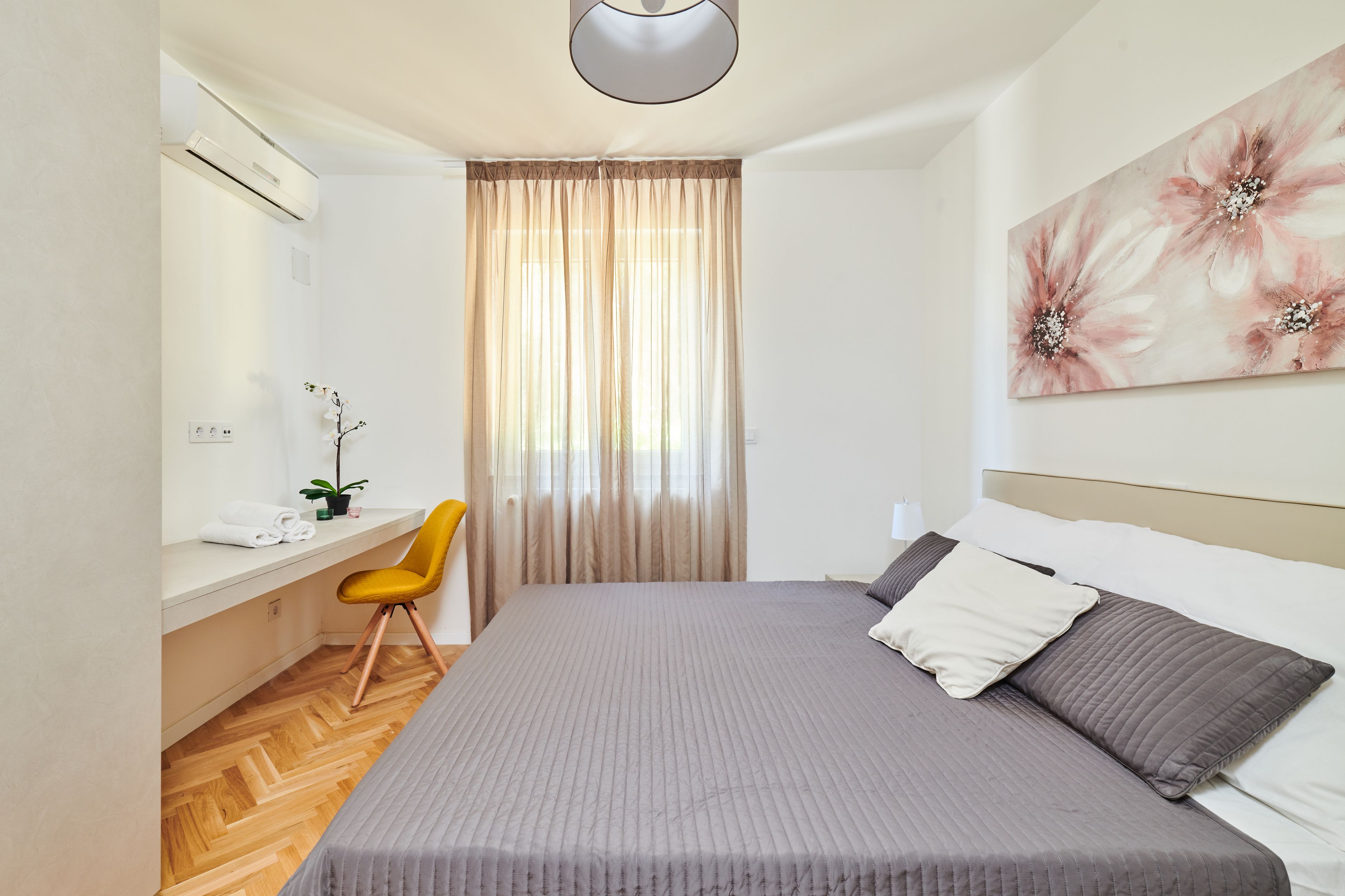 A bedroom with a bed, a desk, a chair, a potted plant, and a painting.