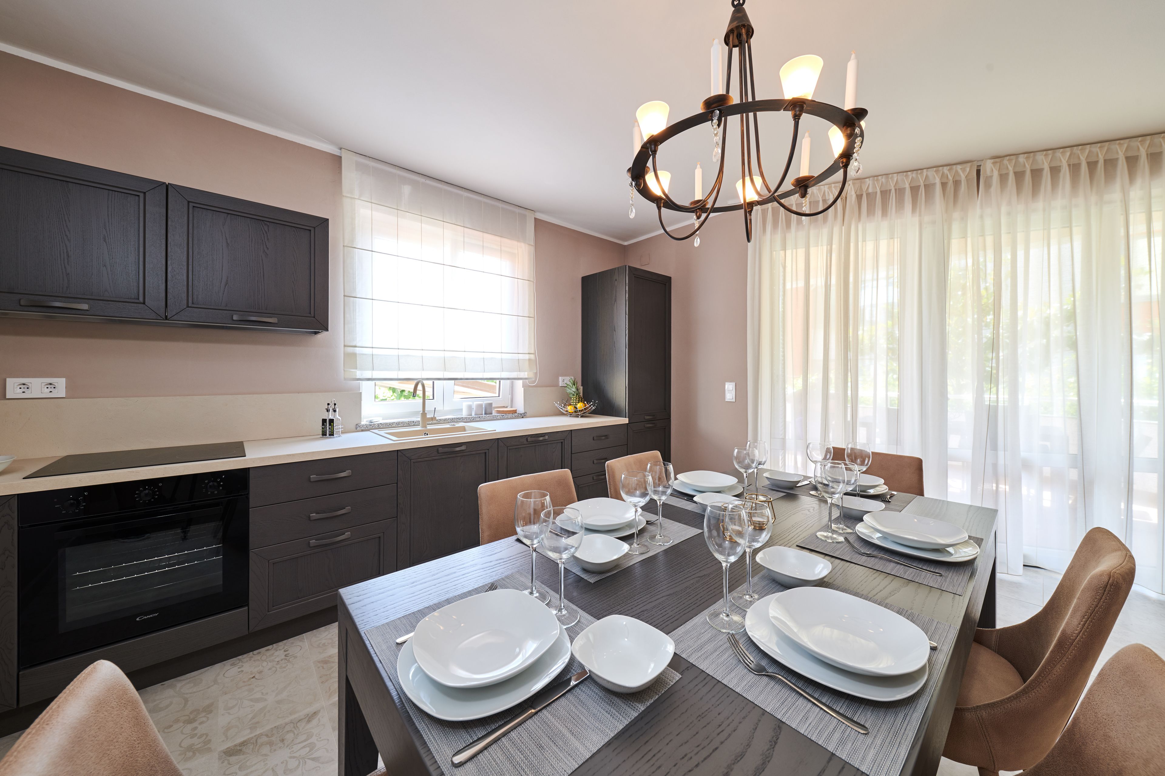 A modern kitchen with dark cabinetry, a built-in oven, and a dining area set for six with a large table, chairs, and a chandelier.