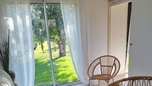 A modern room with a bed, a wicker chair, and a dining set near a large window with a view of a green garden. - La Sellerie (Romagné)