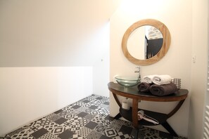 A bathroom with a patterned floor, a round mirror, a glass sink, and towels on a small table. - La Sellerie (Romagné)