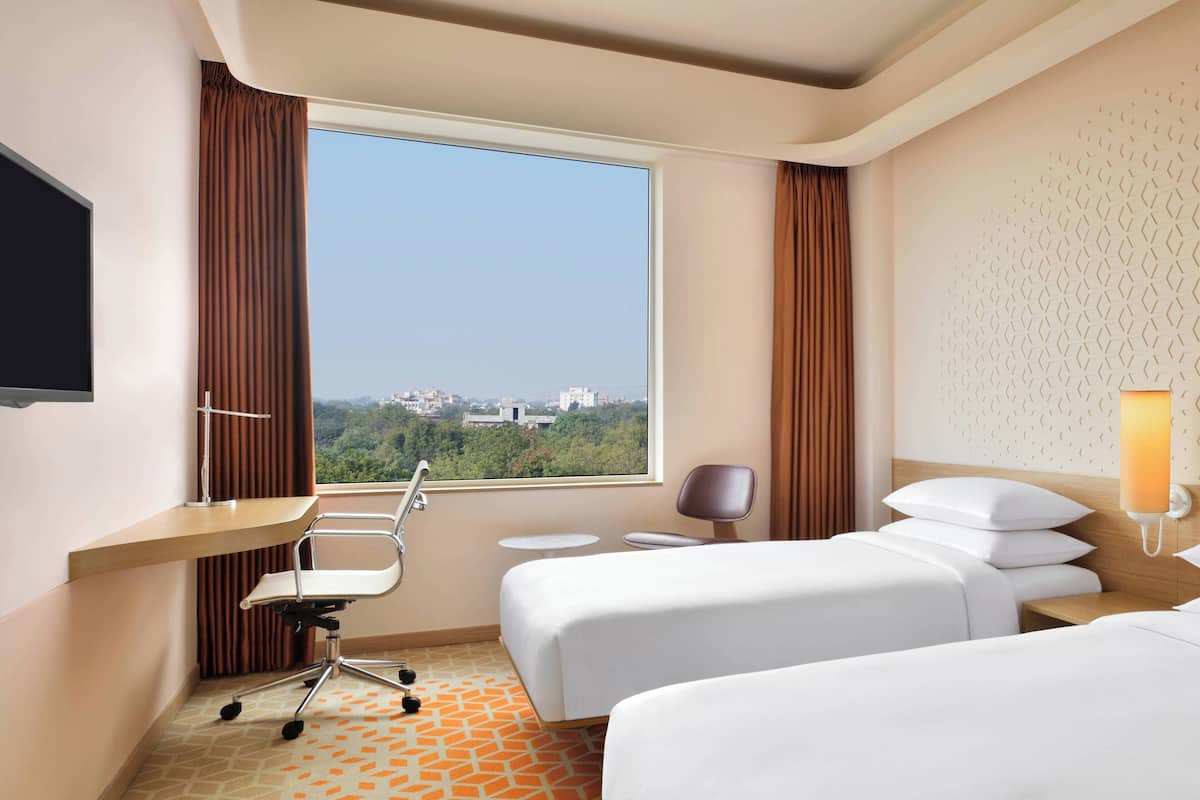 Une chambre d’hôtel avec une grande fenêtre, un bureau, deux lits, une chaise et une lampe.