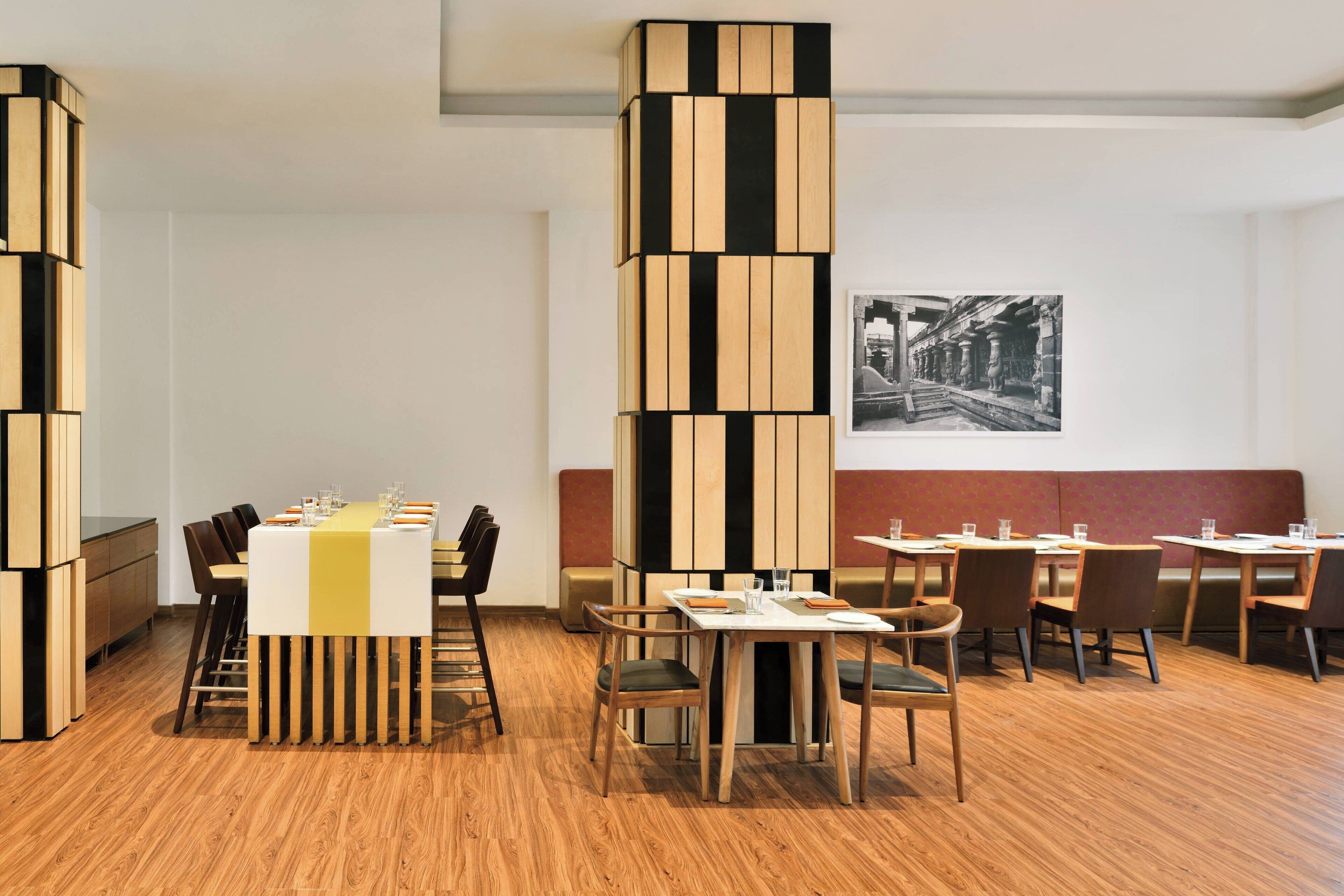 A modern dining area with wooden flooring, a central table with a yellow tablecloth, and a tall column with black and white panels.