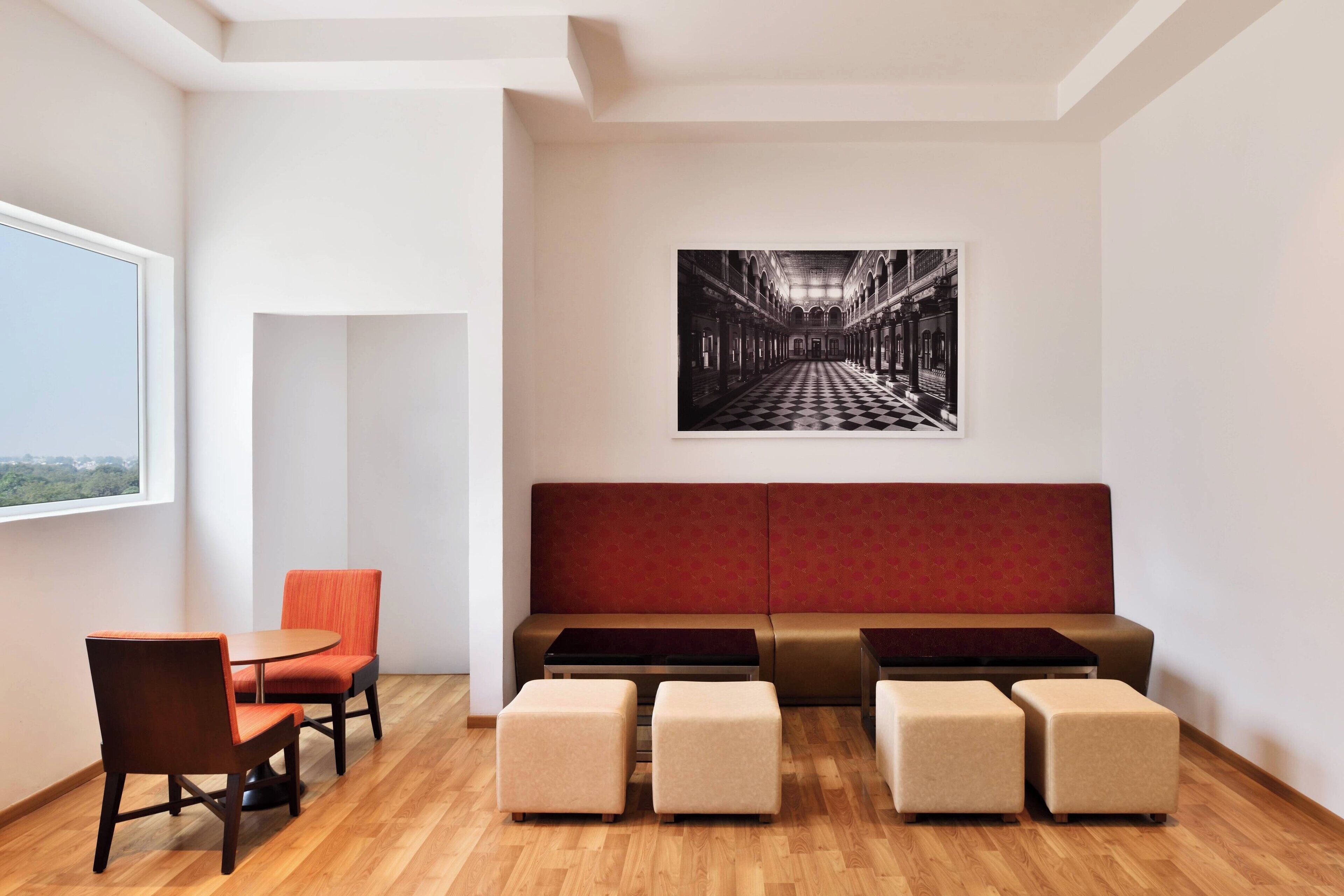 A modern hotel room with a wooden floor, a red sofa, a small table, and a chair. There is a large window on the left side.