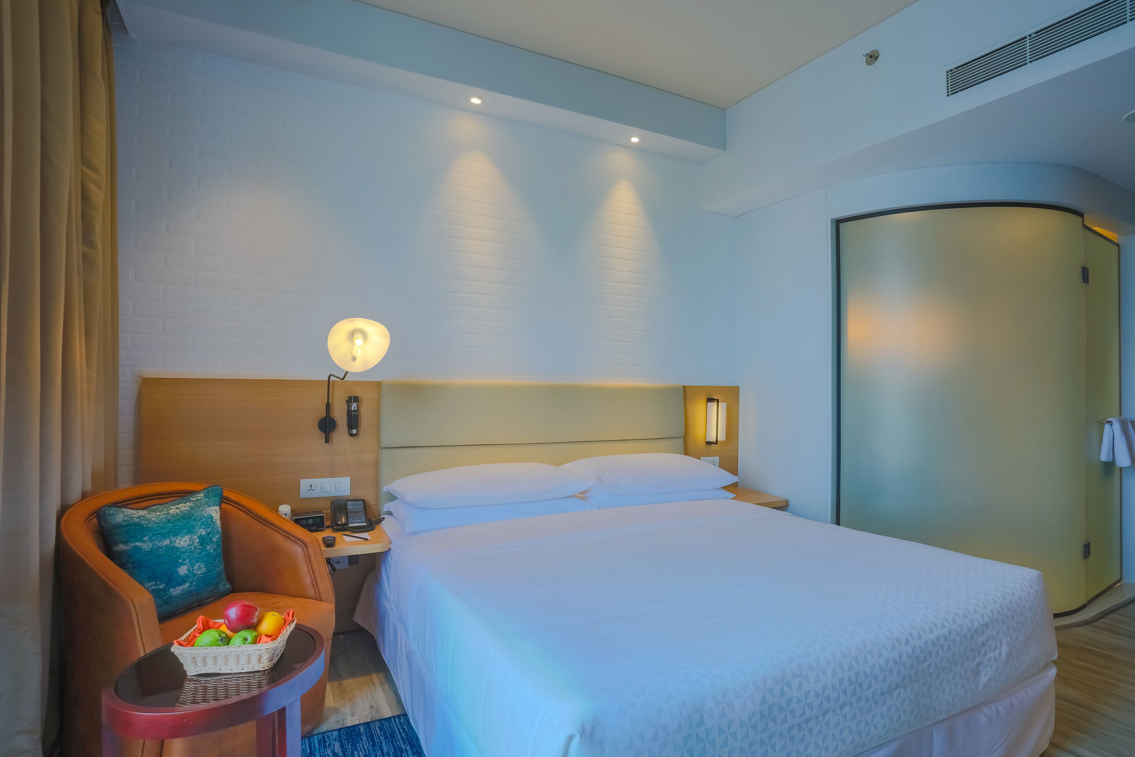 A hotel room with a large bed, a chair with a basket of fruit, a bedside table with a lamp, and a wardrobe.