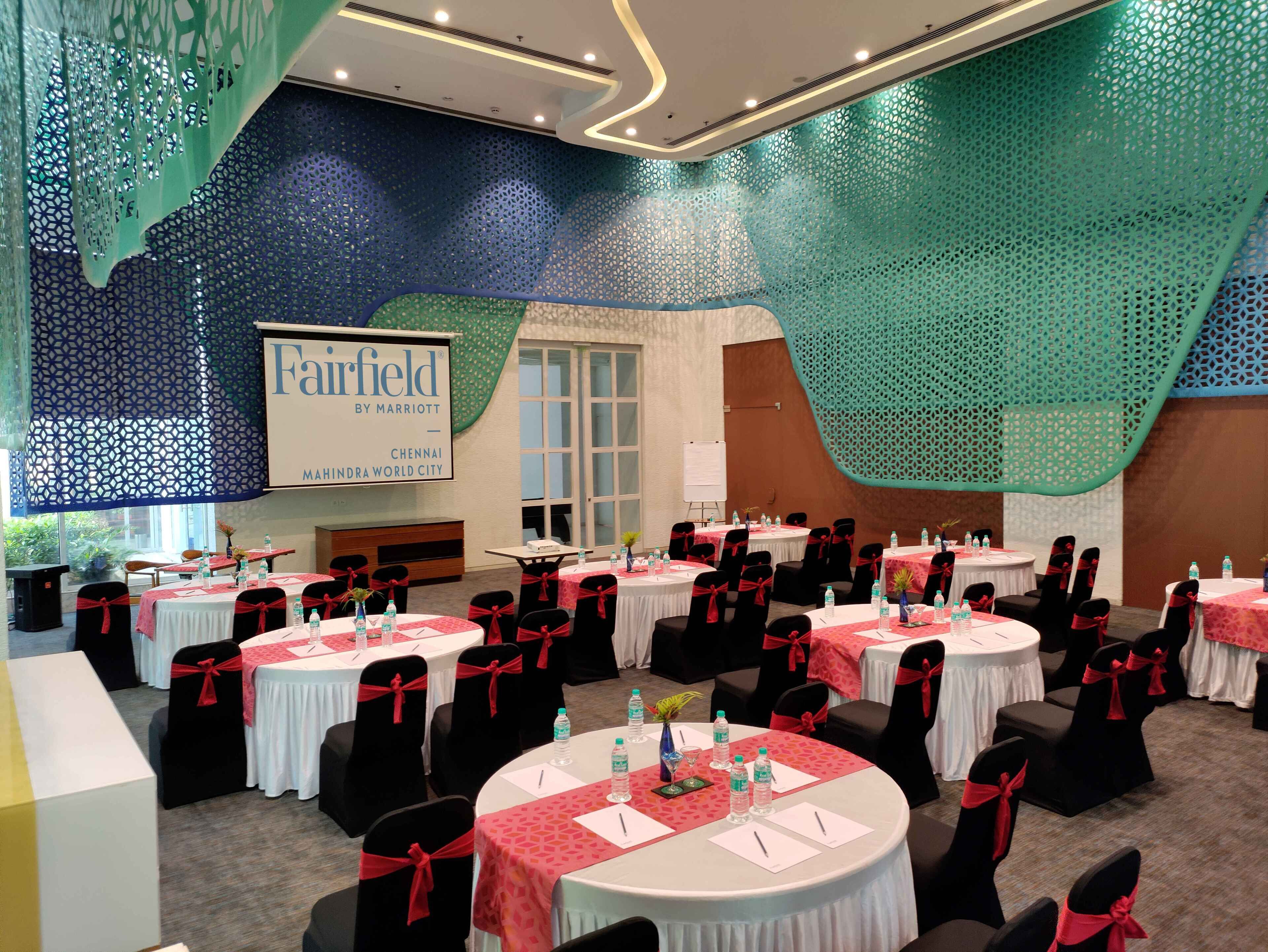 A conference room with round tables, black chairs with red sashes, and a large screen displaying 'Fairfield by Marriott Chennai'.
