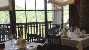 Un espace repas avec des tables préparées pour deux personnes, agrémenté de nappes blanches, de serviettes et de verres à vin. De grandes fenêtres offrent une vue sur un paysage verdoyant et luxuriant.