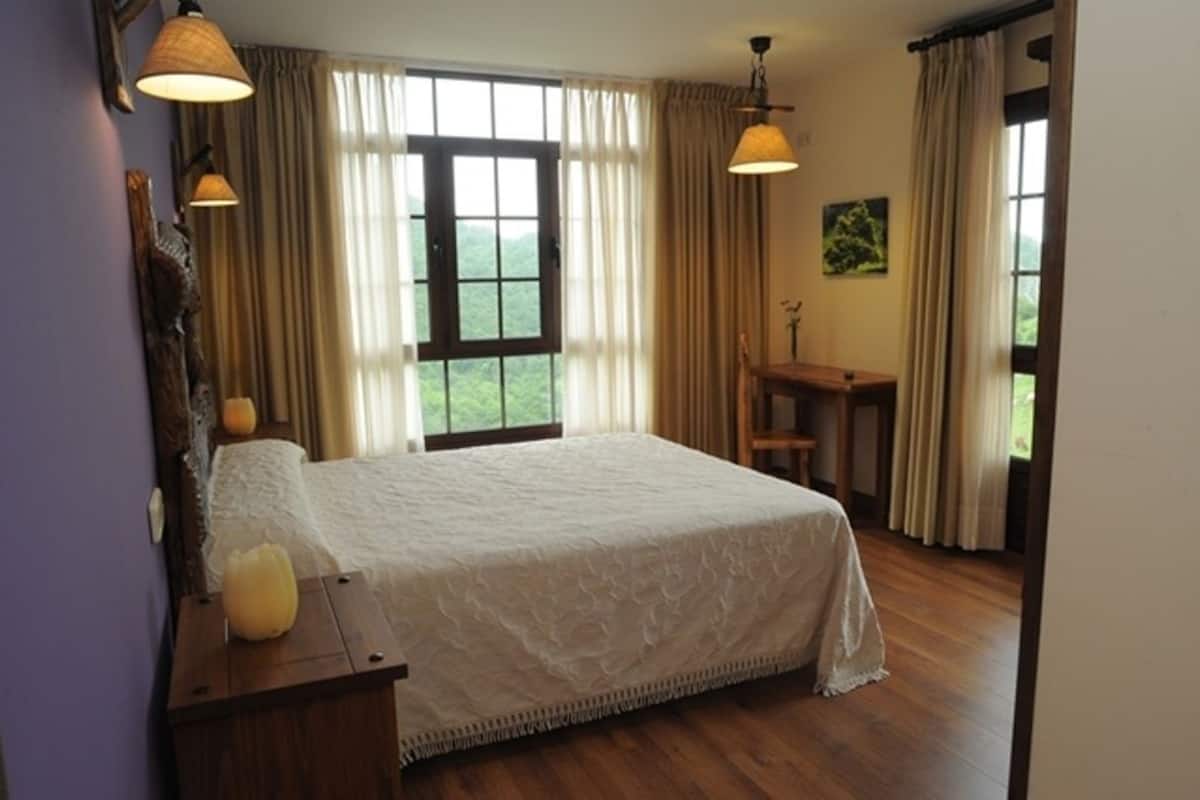 Een slaapkamer met een groot bed, houten vloeren, een bureau en uitzicht op groen buiten het raam.
