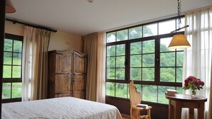 Une chambre avec une grande fenêtre, une armoire en bois, un lit recouvert d’une courtepointe blanche, une chaise en bois et une petite table sur laquelle est posé un vase de fleurs.
