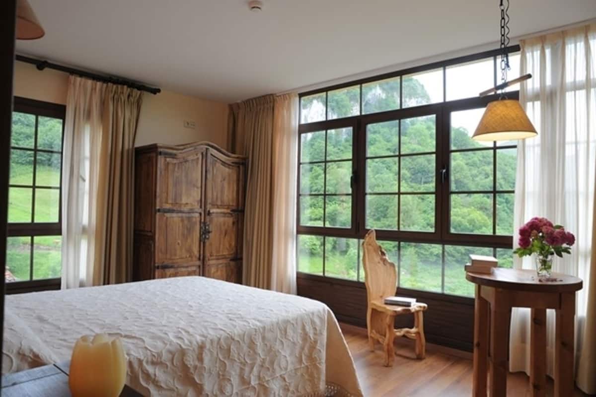 Een slaapkamer met een groot raam, een houten garderobe, een bed met een witte dekbedovertrek, een houten stoel en een klein tafeltje met een bloevas.