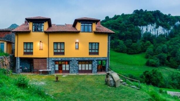 Un bâtiment jaune à deux étages, doté d’une façade en pierre, qui se dresse au milieu d’un paysage verdoyant, avec une falaise en arrière-plan.