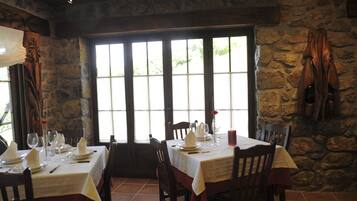 A dining area with stone walls and large windows, tables set with white tablecloths and red napkins, and wooden chairs.