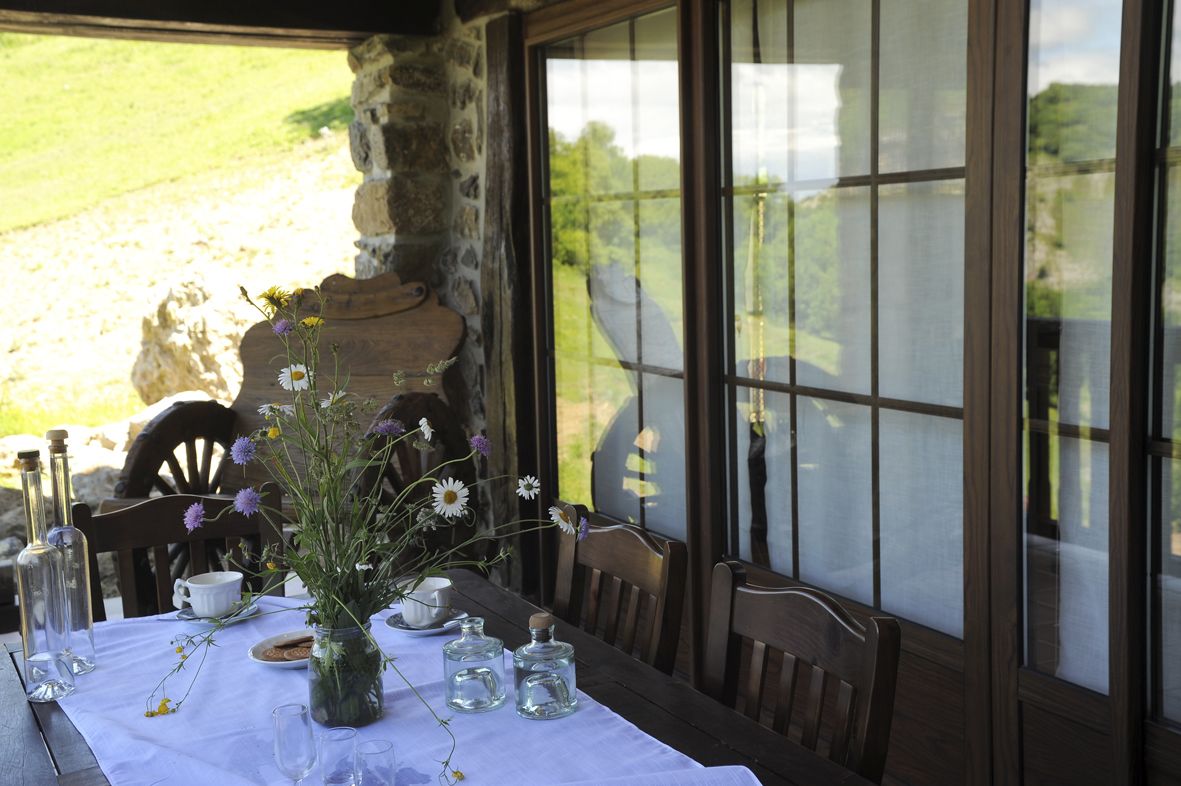 A table set with a vase of flowers, a cup, and a plate, with a stone wall and a window in the background.