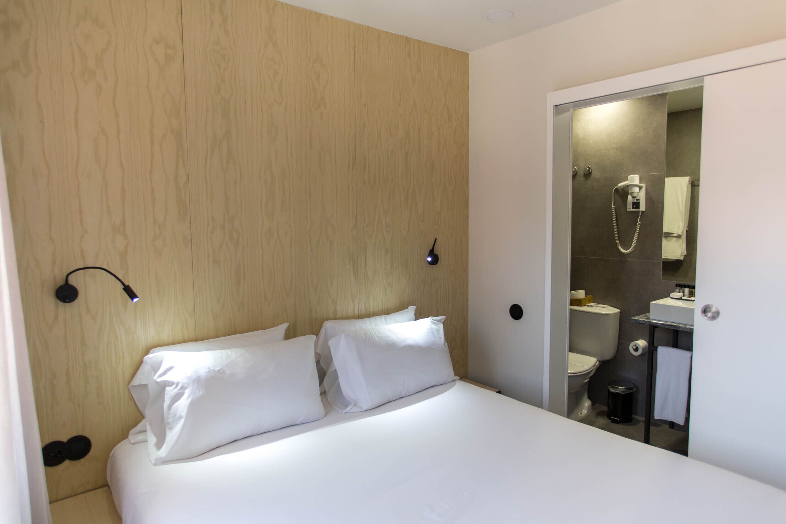 Habitación de hotel con cabecera de madera, cama con ropa blanca, lámpara de noche y vista al baño a través de una puerta abierta.