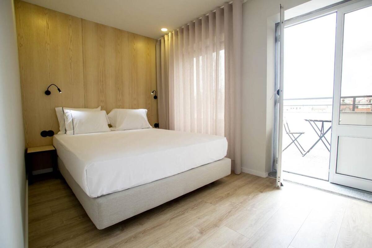 A modern bedroom with a large bed, wooden panel walls, a small bedside table, and a view of the outside balcony.