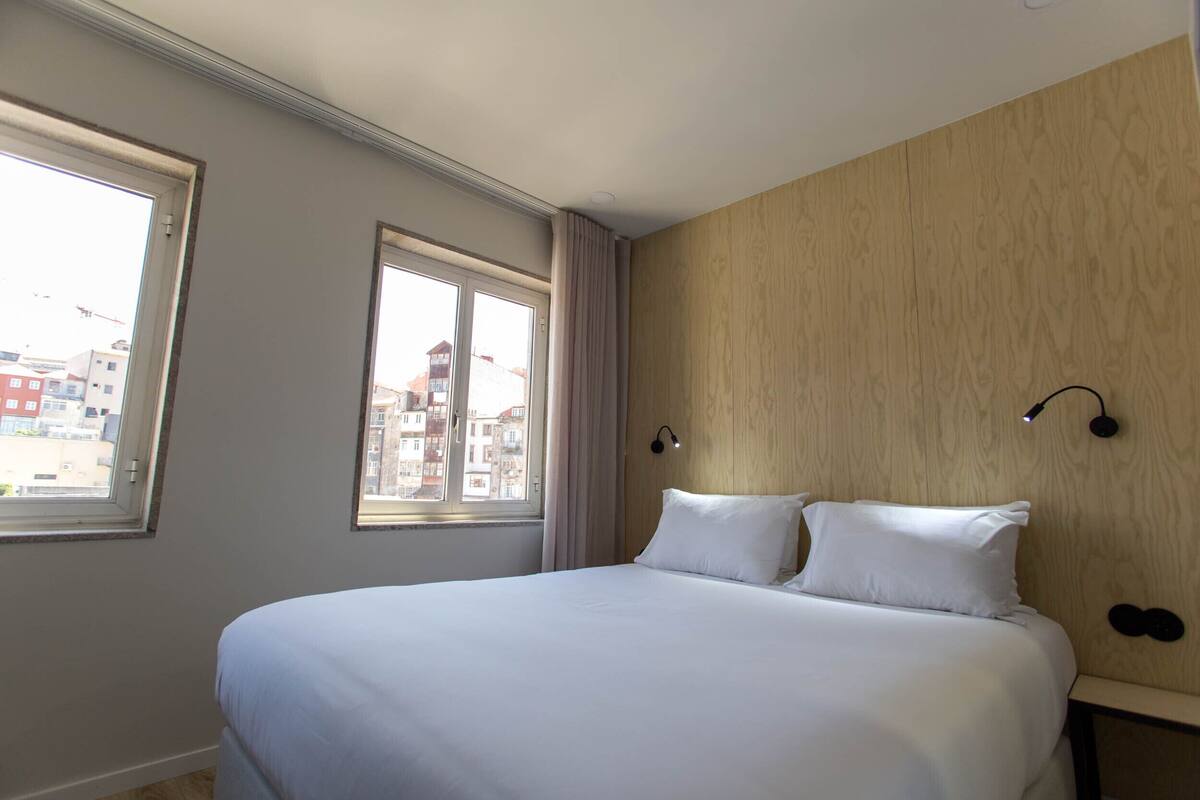 A hotel room with a large bed, two windows, and a view of buildings outside.
