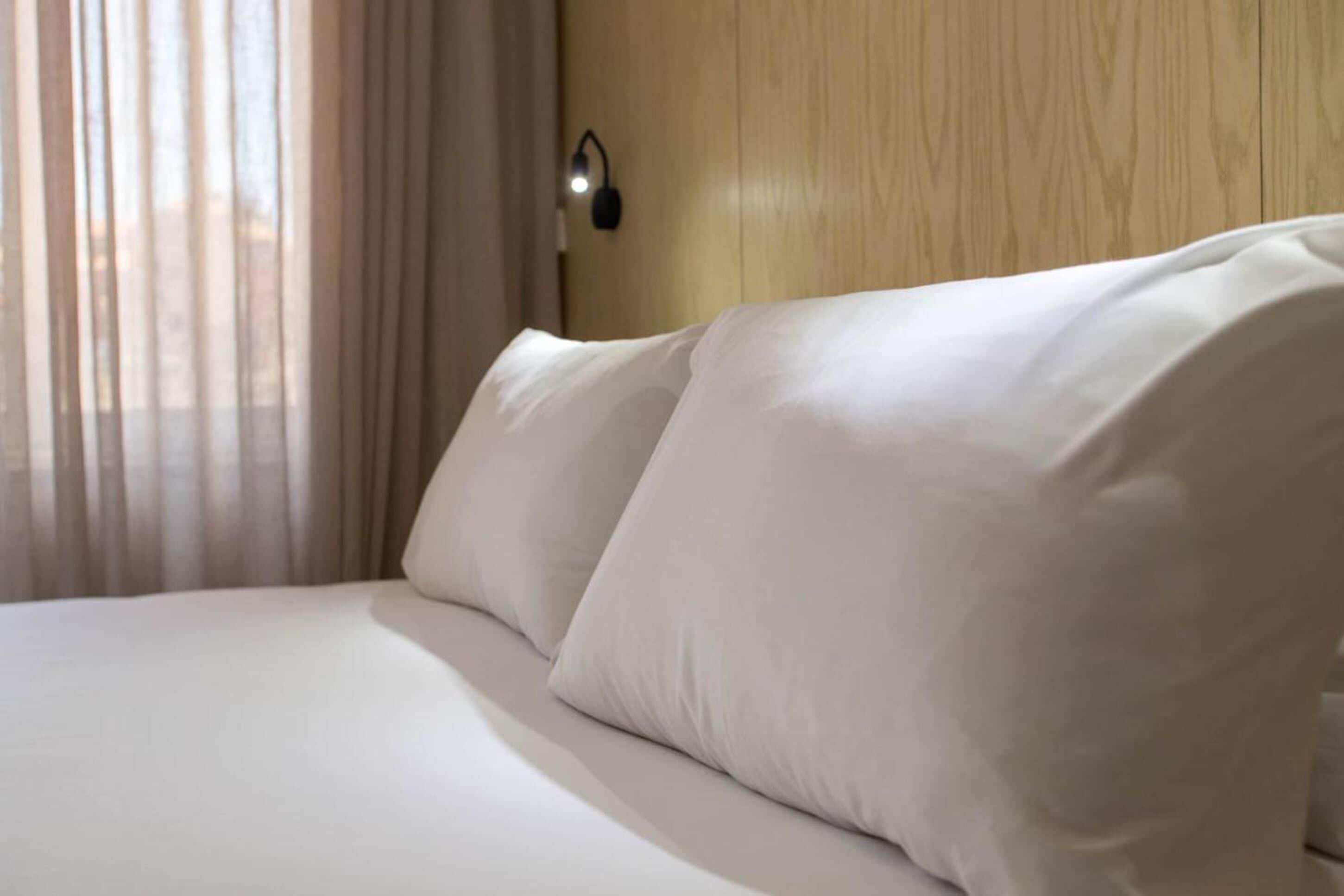 A neatly made bed with white linens and pillows, a wooden headboard, and a wall-mounted light fixture.