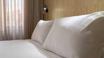 A neatly made bed with white linens and pillows, a wooden headboard, and a wall-mounted light fixture.