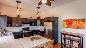 Une cuisine avec des meubles en bois foncé, un réfrigérateur en acier inoxydable, une cuisinière à gaz, un ventilateur de plafond et un coin repas avec des chaises et une table en osier.