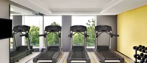 A gym with four treadmills aligned in a row, a mirror on the left, and a rack of dumbbells on the right.